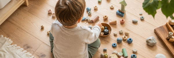 Kind beim Spielen mit kleinen bunten Spielzeugteilen und Holzperlen auf einem hellen Holzboden — symbolisch für das Neugierverhalten von Kleinkindern