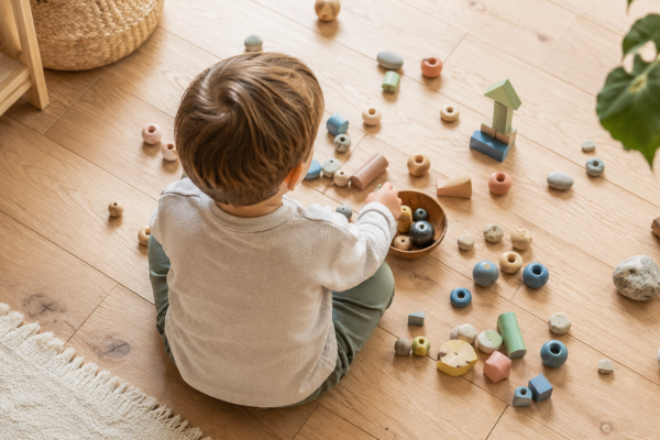 Kind beim Spielen mit kleinen bunten Spielzeugteilen und Holzperlen auf einem hellen Holzboden — symbolisch für das Neugierverhalten von Kleinkindern